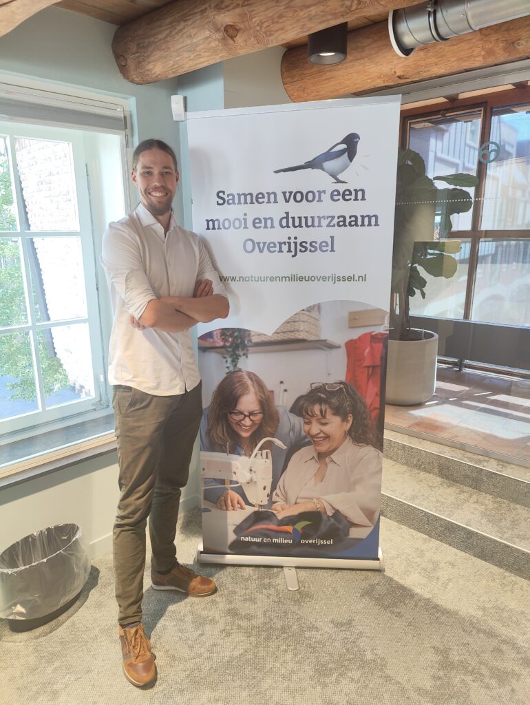 Collega Jurian poseert naast een banner met de slogan van Natuur en Milieu Overijssel. Hij staat met zijn handen over elkaar, breed lachend bij het raam.